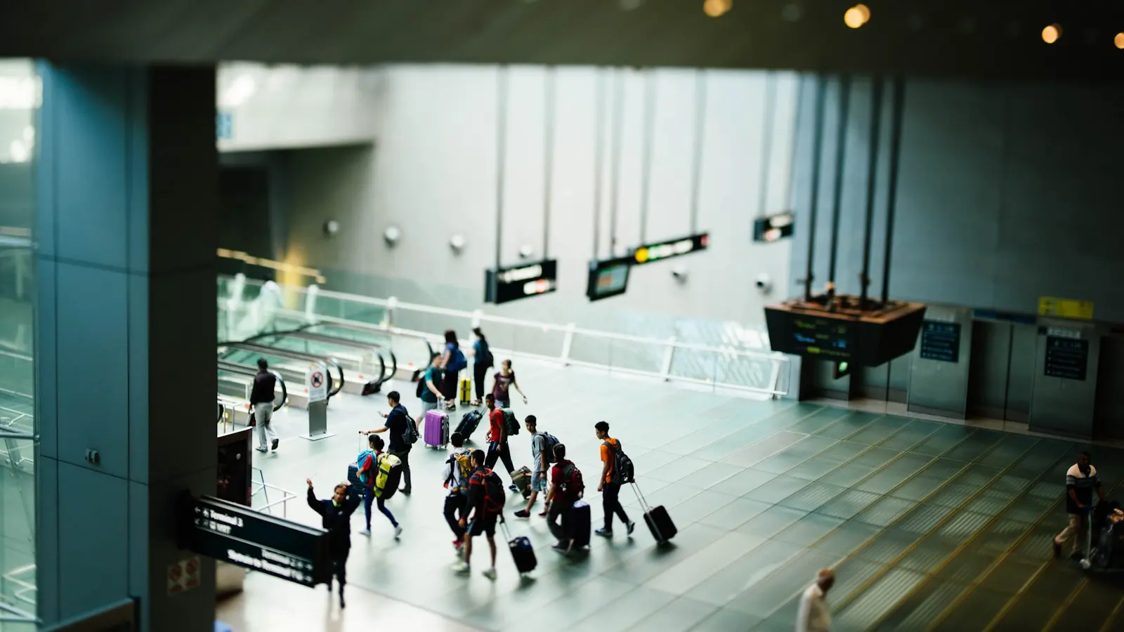 Travelers wheeling carry-on suitcases through an airport terminal past departure signage