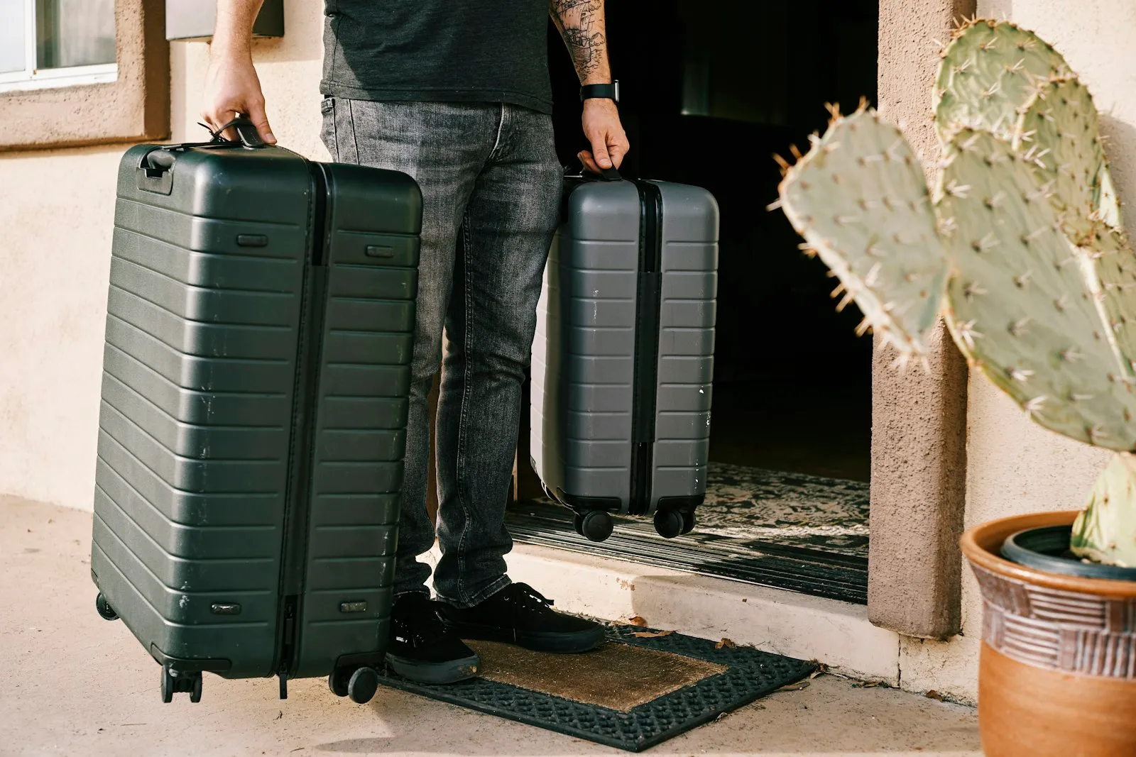 A person carrying two ribbed hardside suitcases, a large checked bag and a carry-on, out a doorway