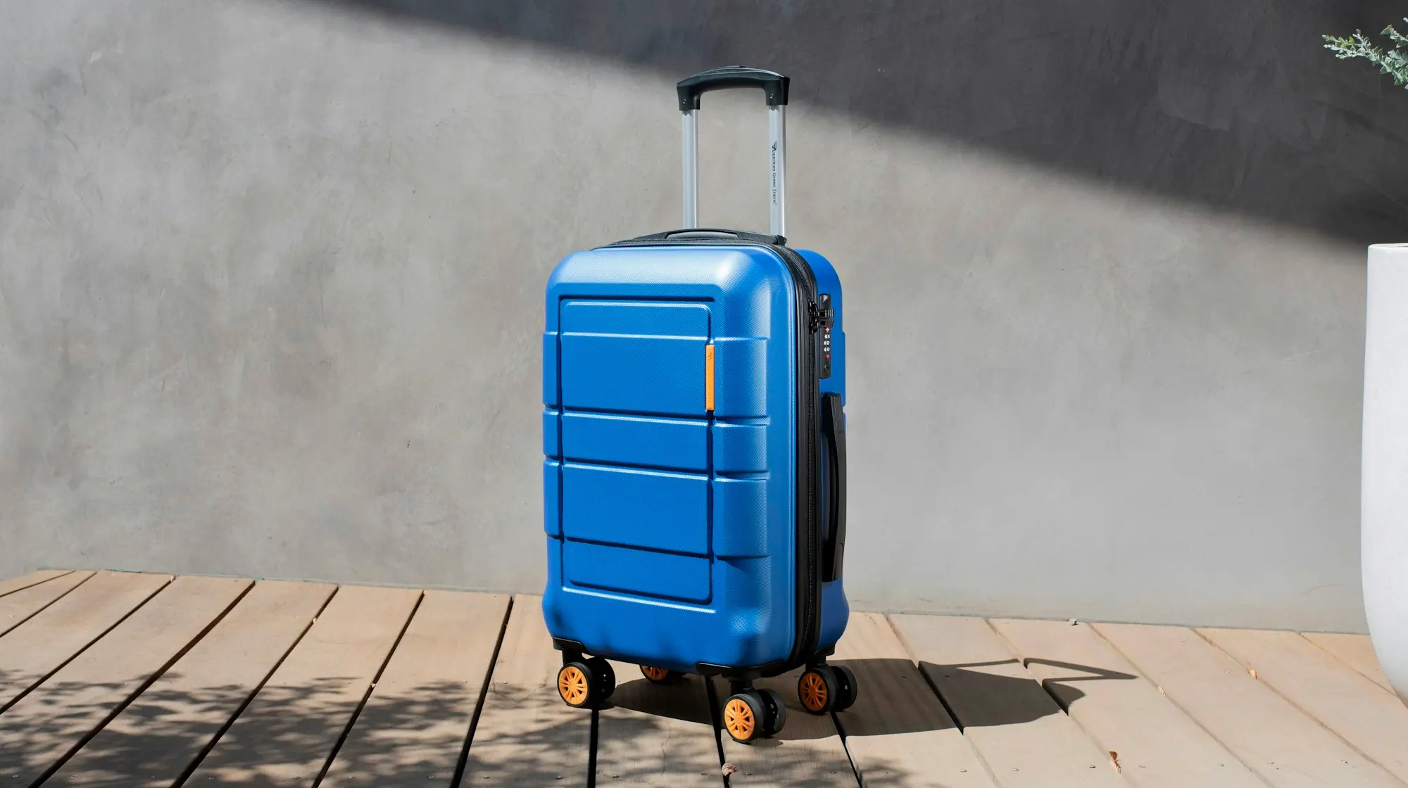 A blue hardside carry-on suitcase with its telescoping handle up and orange spinner wheels, standing on a wooden deck in warm sunlight