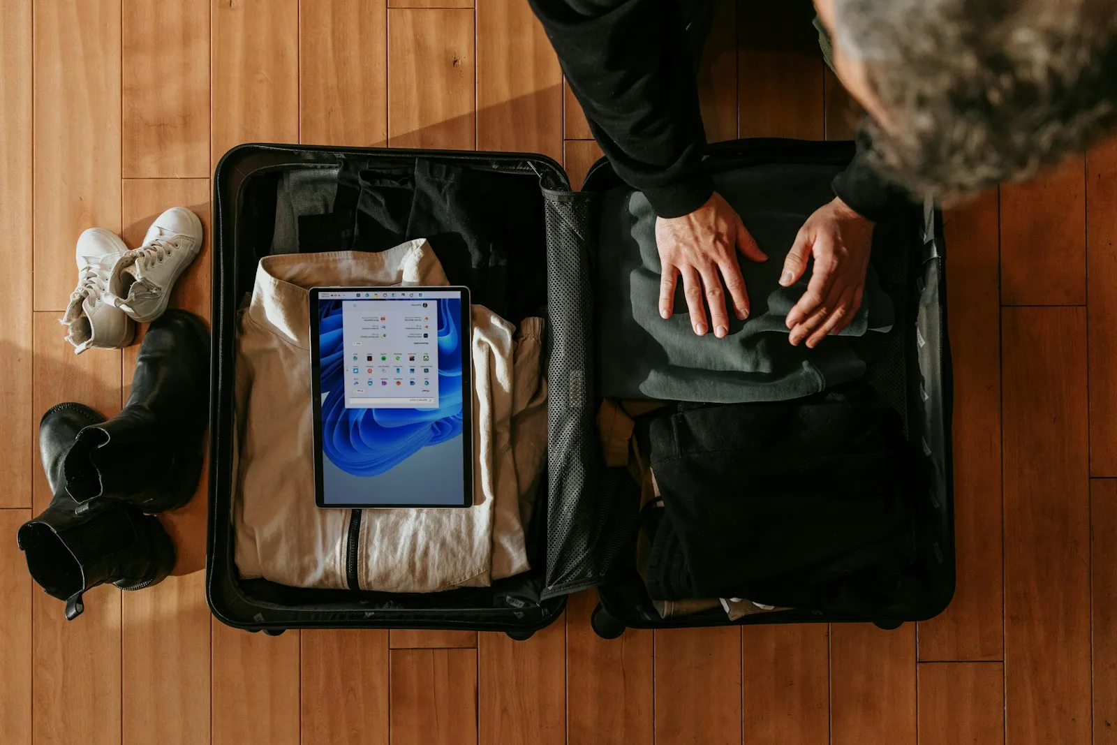 An overhead view of a traveler pressing folded clothes flat into an open carry-on suitcase on a wooden floor