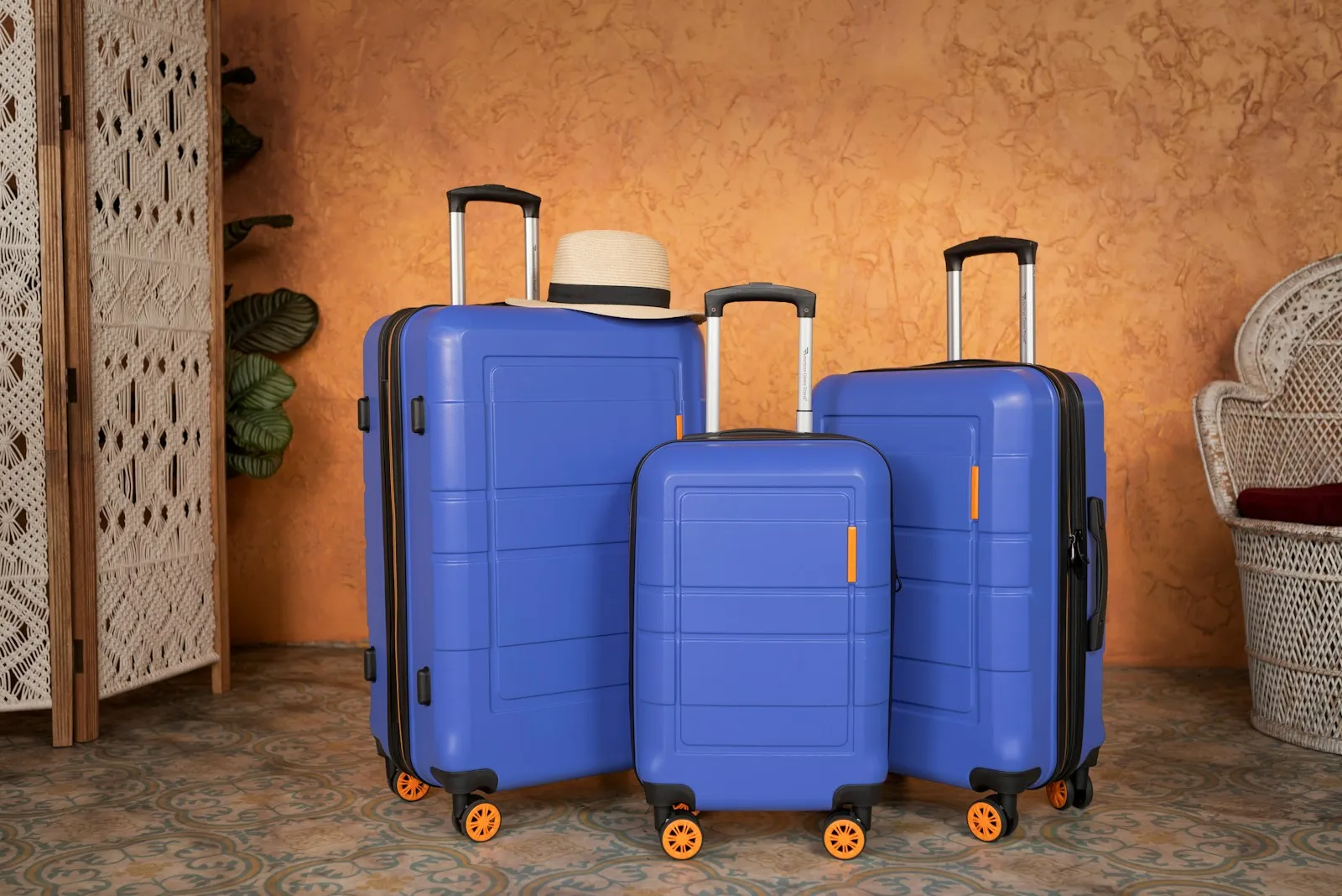 A matching three-piece set of blue hardside suitcases with orange spinner wheels standing against an ochre wall