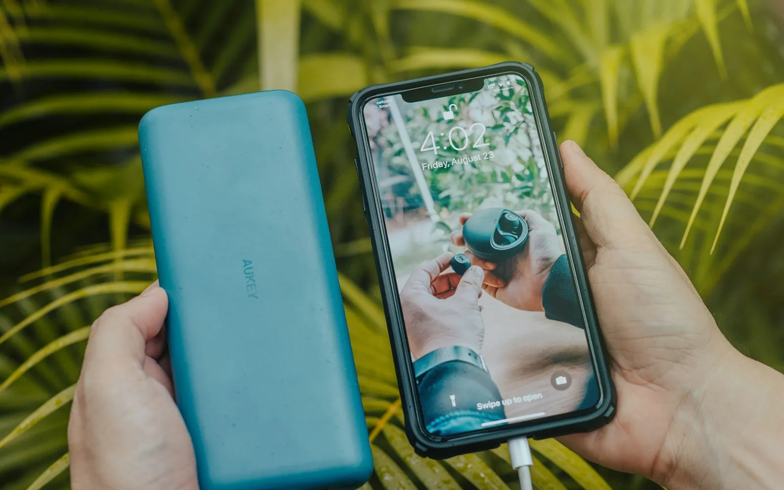 A hand holding a teal portable power bank beside a smartphone charging, against green palm leaves