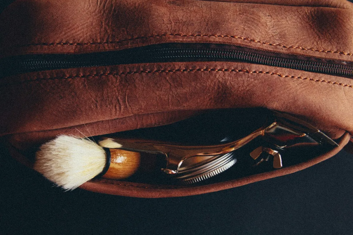 A brown leather dopp kit unzipped to show a shaving brush and a gold safety razor tucked inside