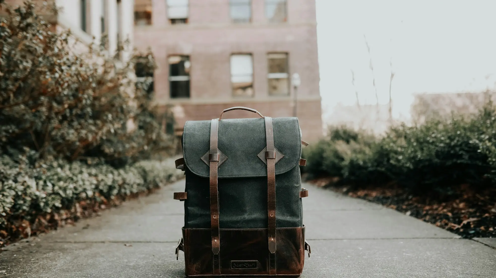 A waxed-canvas and leather travel backpack standing on a city sidewalk with a brick building behind it