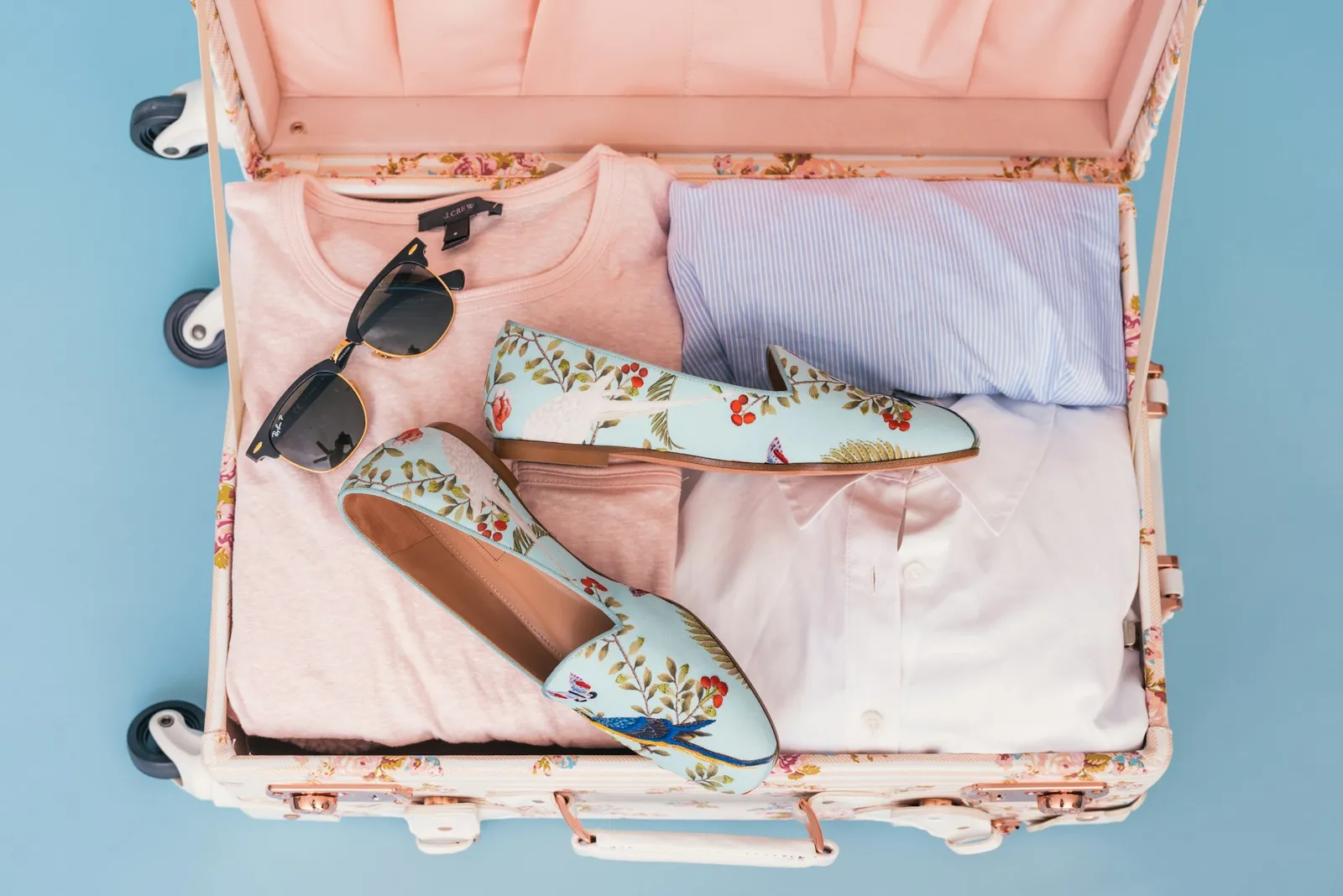 An open suitcase packed with folded clothes, shoes, and sunglasses on a blue background