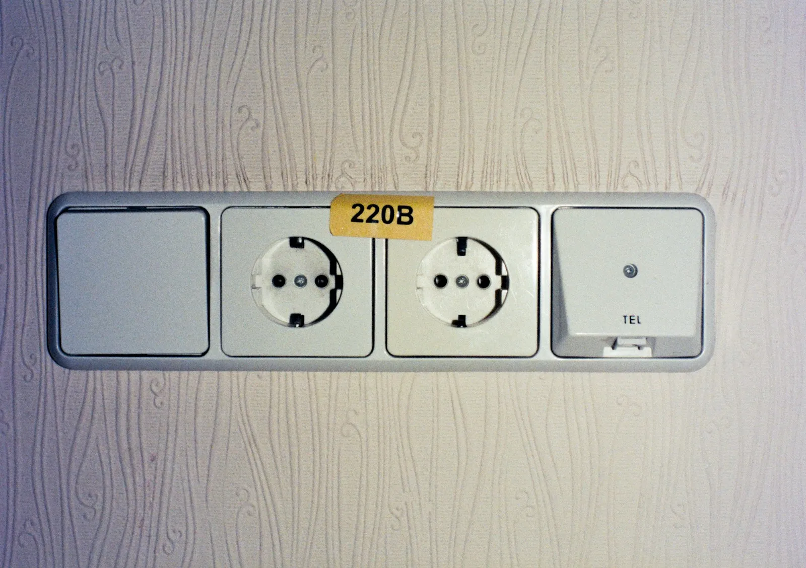 A row of European wall sockets with a yellow 220V label on a wallpapered hotel wall