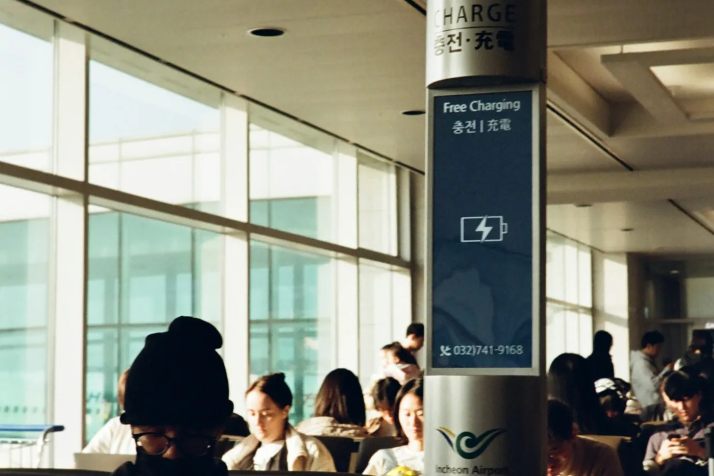 The Free Charging station at Incheon Airport, a lit pillar with a battery icon, travelers seated around it in sunlight
