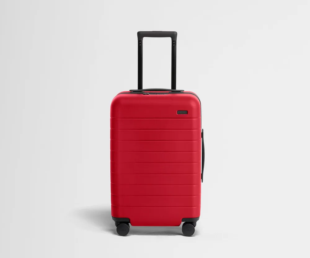The Away Carry-On hardside spinner suitcase in cherry red, shown at an angle with its four spinner wheels visible
