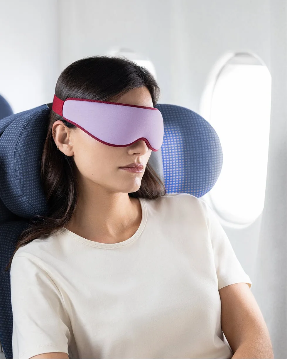 A traveler on a plane wearing the lilac and red Ostrichpillow contoured eye mask beside a window