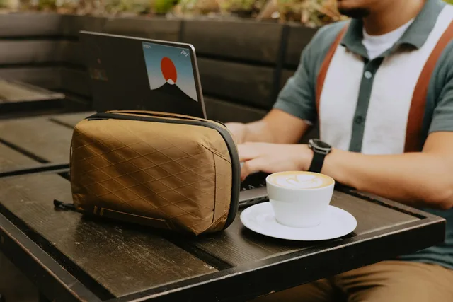 The Peak Design Tech Pouch in coyote tan, a boxy zippered tech organizer with its origami interior folded open to show cables and chargers in labeled slots