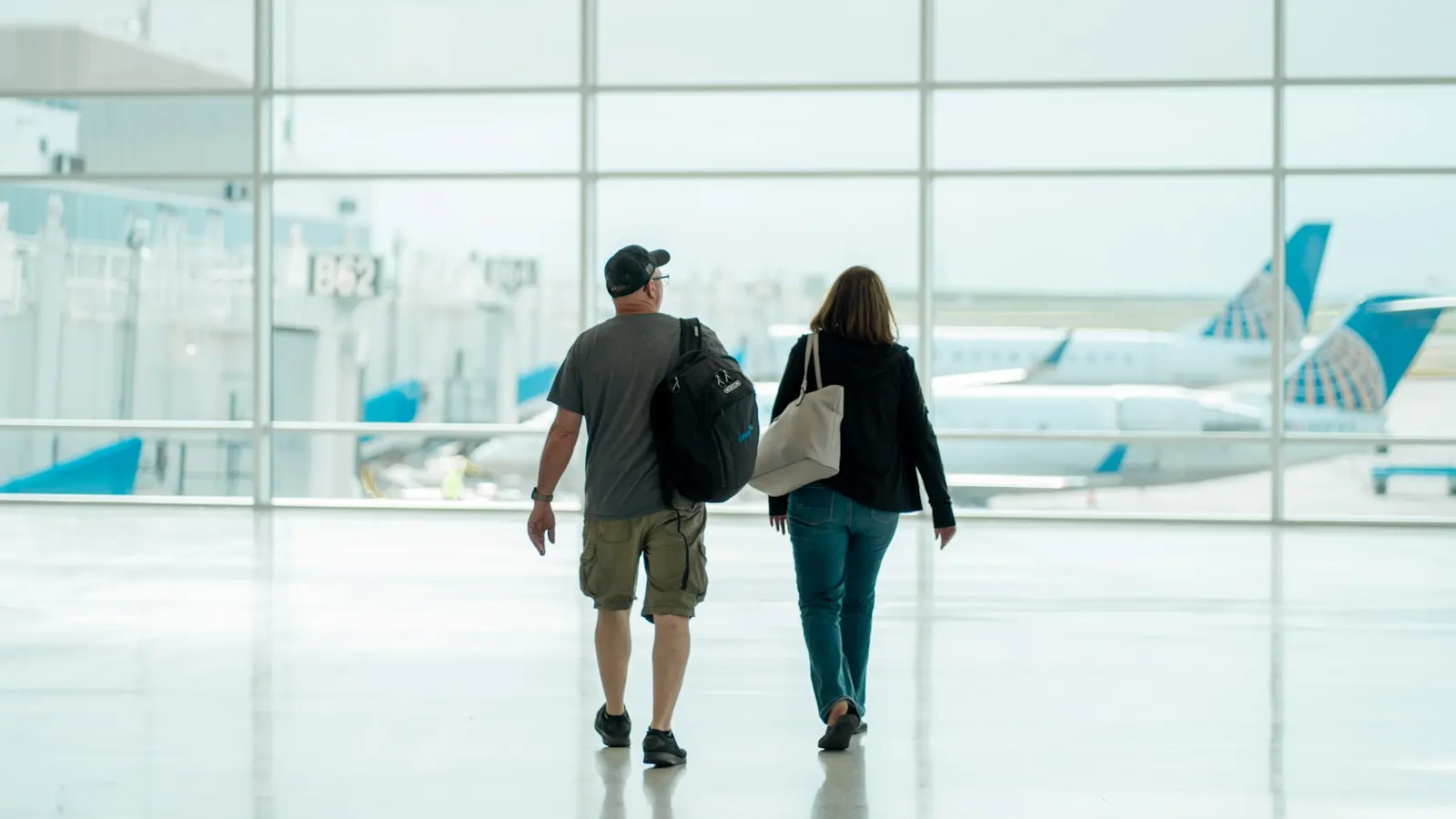 A traveler wearing a backpack walking through an airport terminal with parked planes visible through the windows