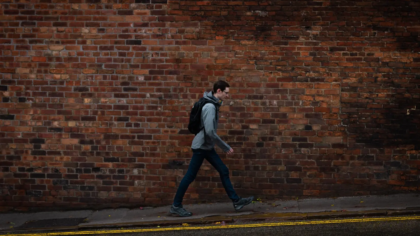 A traveler walking along a city street wearing a backpack, seen from the side against a brick wall